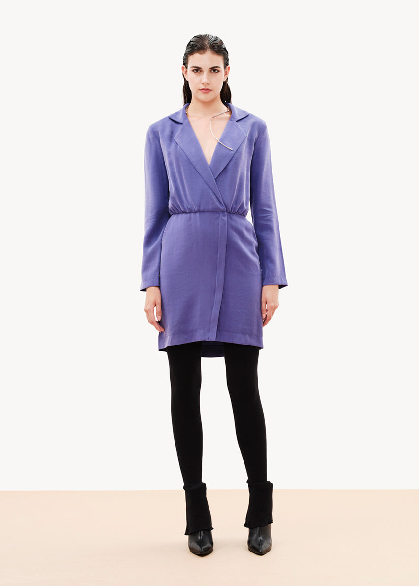 A woman standing wearing a purple draped blazer dress with a lapel collar, long sleeves, and a front zipper.