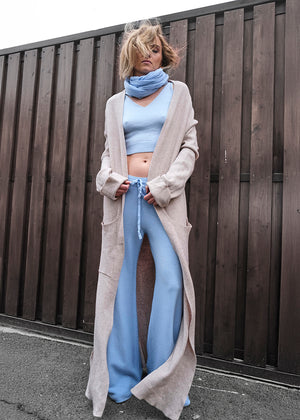 Person wearing a long beige cardigan, light blue top, and pants against a wooden fence.