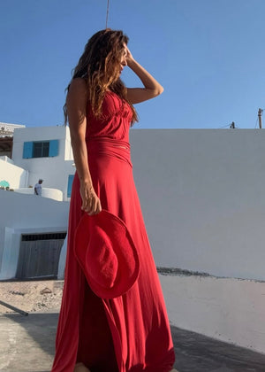 Woman in a red dress holding a red hat against a white building and blue sky.