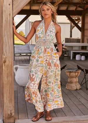 A woman standing on a porch wearing a beige and multicolor floral jumpsuit with a halter neckline and wide-leg design.