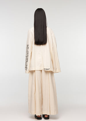 A model wearing a beige oversized jacket with an open collar, featuring four front pockets and closures with buttons. The jacket has a relaxed fit and the model is also wearing light-colored pants.