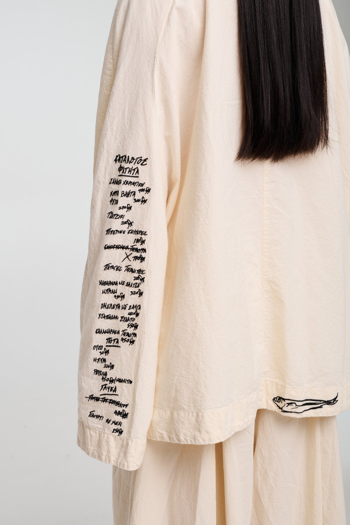 A model wearing a beige oversized jacket with an open collar, featuring four front pockets and closures with buttons. The jacket has a relaxed fit and the model is also wearing light-colored pants.