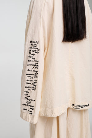 A model wearing a beige oversized jacket with an open collar, featuring four front pockets and closures with buttons. The jacket has a relaxed fit and the model is also wearing light-colored pants.