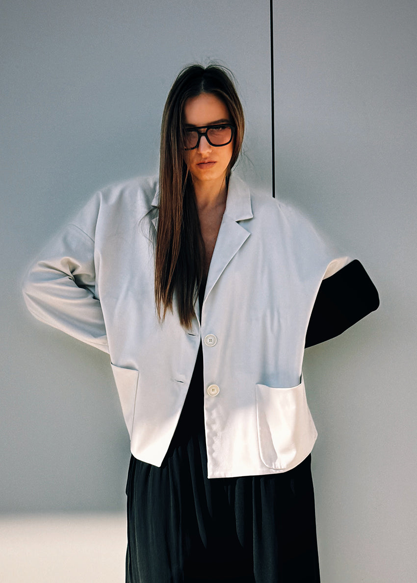 A woman wearing a white short-medium jacket with a lapel collar and black long sleeves, paired with a black wide-legged pant and sunglasses.