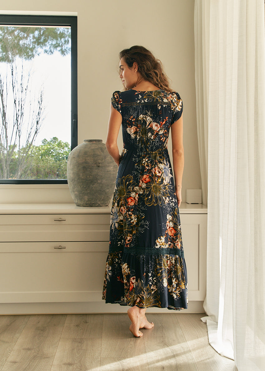 A woman standing in an elegant pose wearing a long floral print dress with a V-neckline and short sleeves.