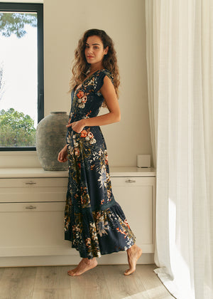 A woman standing in an elegant pose wearing a long floral print dress with a V-neckline and short sleeves.