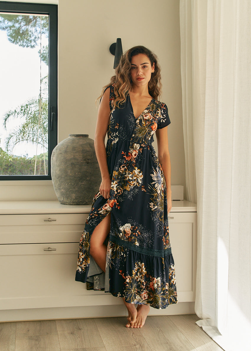 A woman standing in an elegant pose wearing a long floral print dress with a V-neckline and short sleeves.
