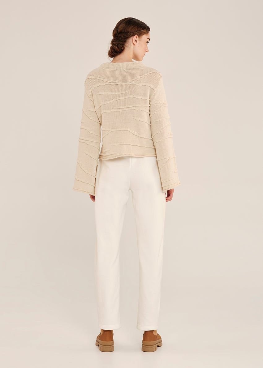 Person wearing a beige sweater and white pants on a plain background