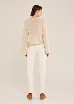 Person wearing a beige sweater and white pants on a plain background