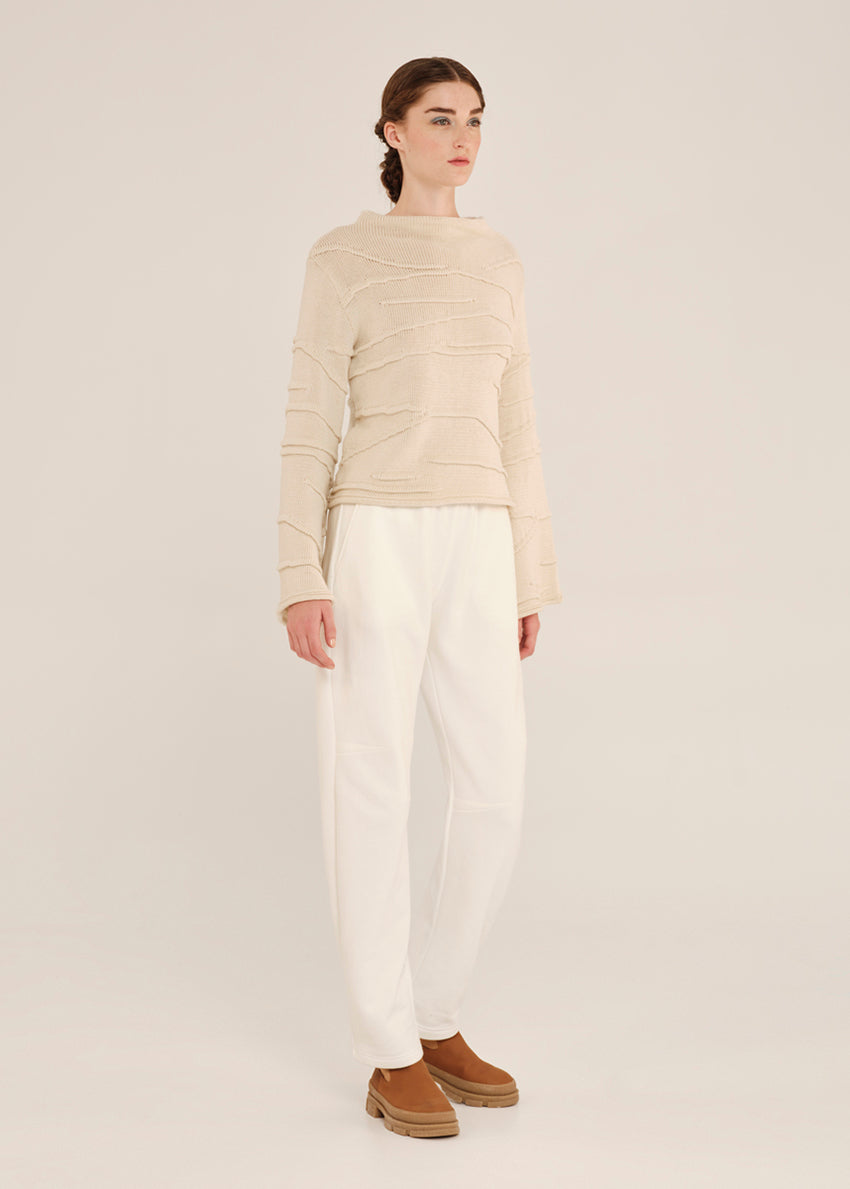 Person wearing a beige sweater and white pants on a plain background