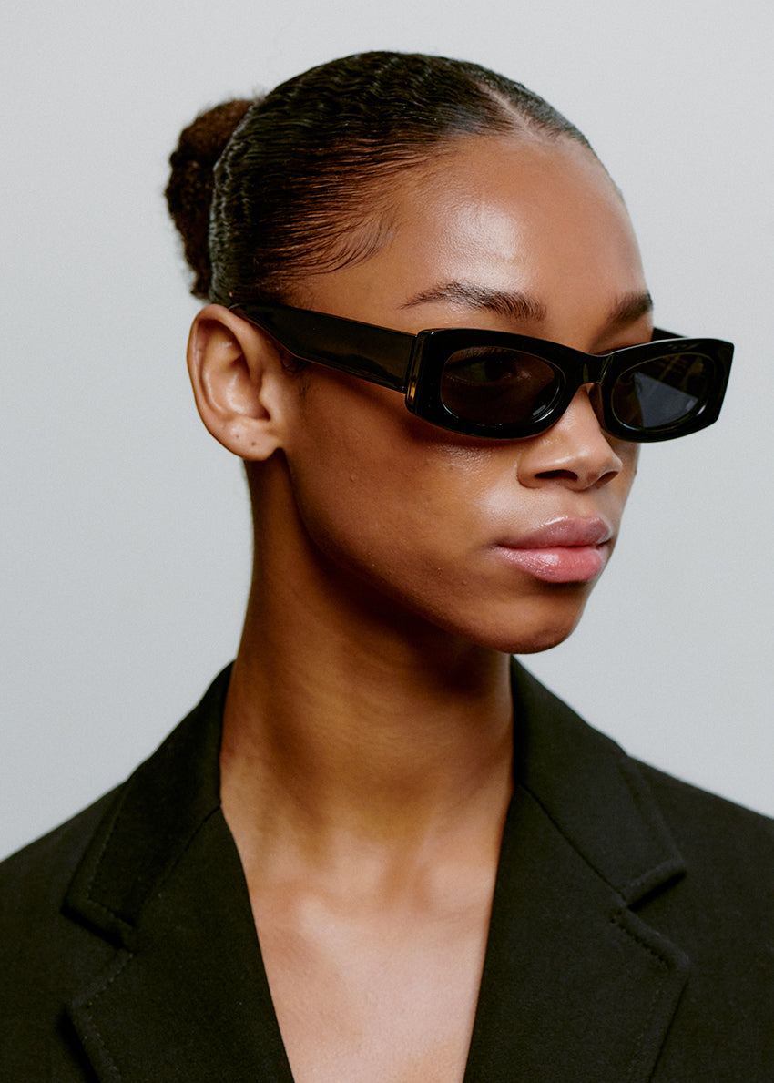 Woman wearing black sunglasses and a black blazer against a gray background
