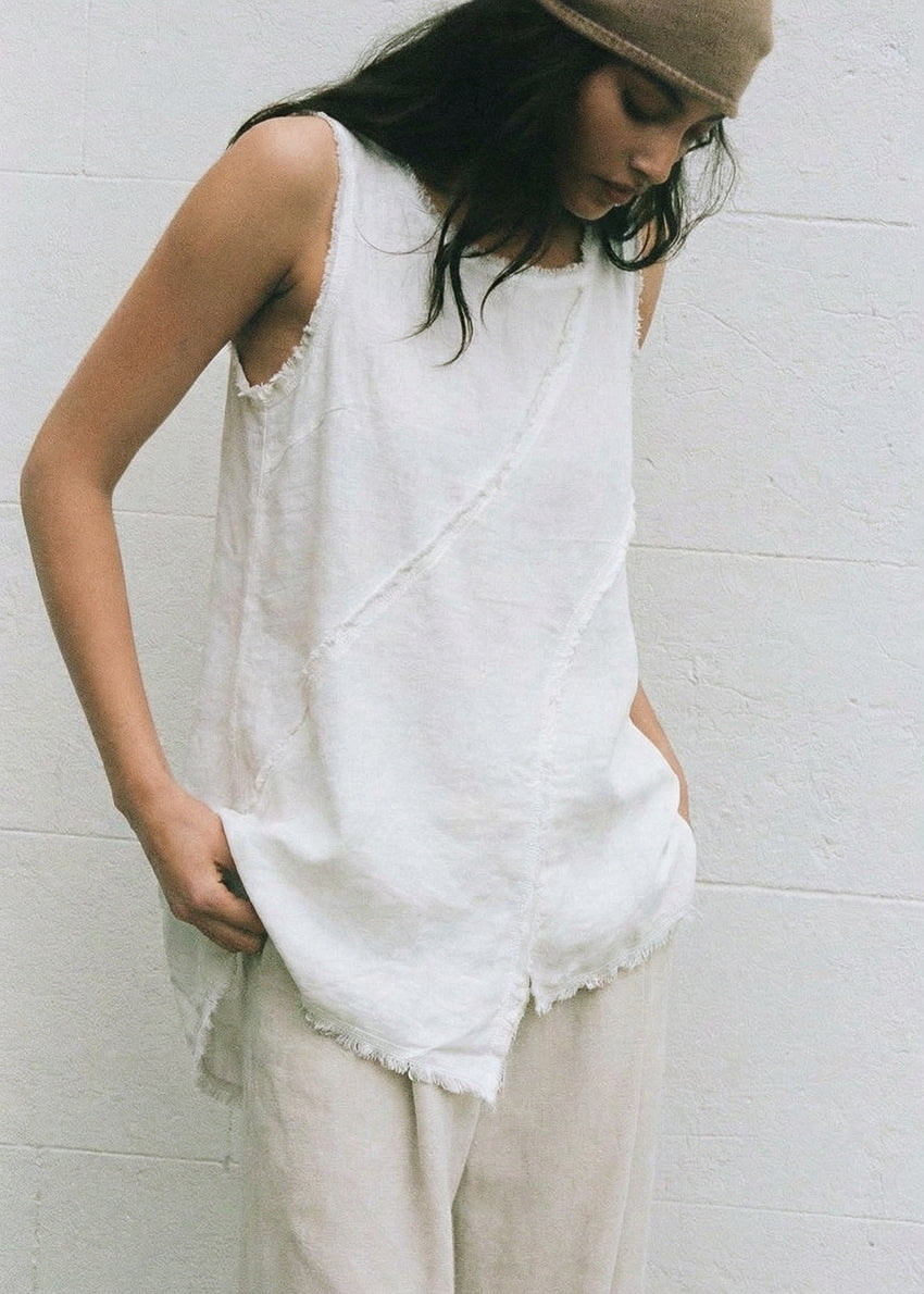 Woman wearing a white sleeveless top and beige pants against a white wall.
