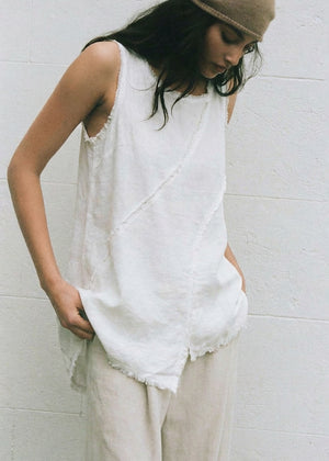 Woman wearing a white sleeveless top and beige pants against a white wall.