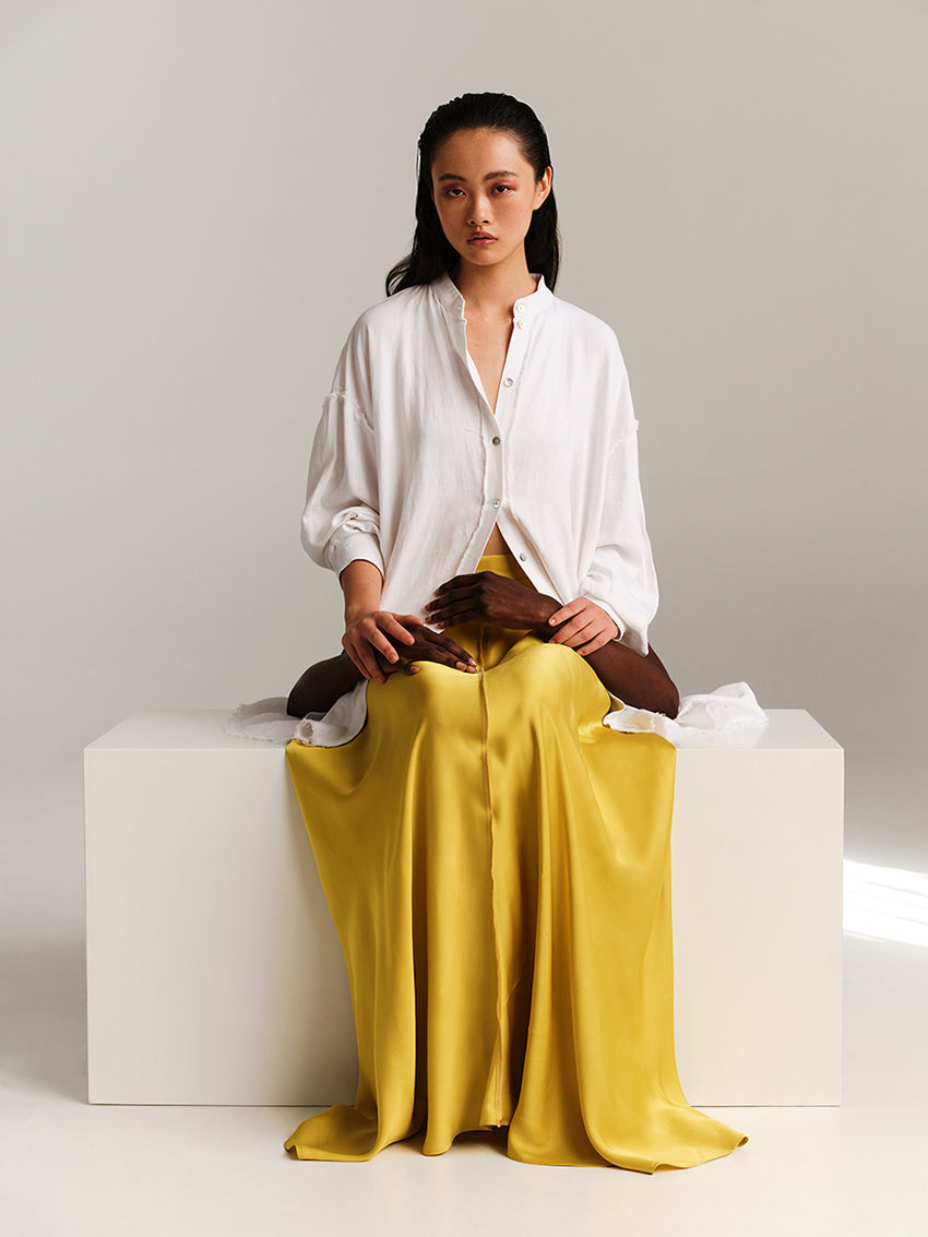 Woman wearing a white shirt and yellow skirt sitting on a white cube against a gray background