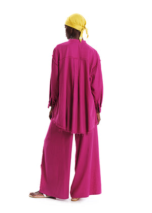 A model wearing a magenta oversized linen and bamboo blend shirt with fringes at the seams, paired with matching pants.