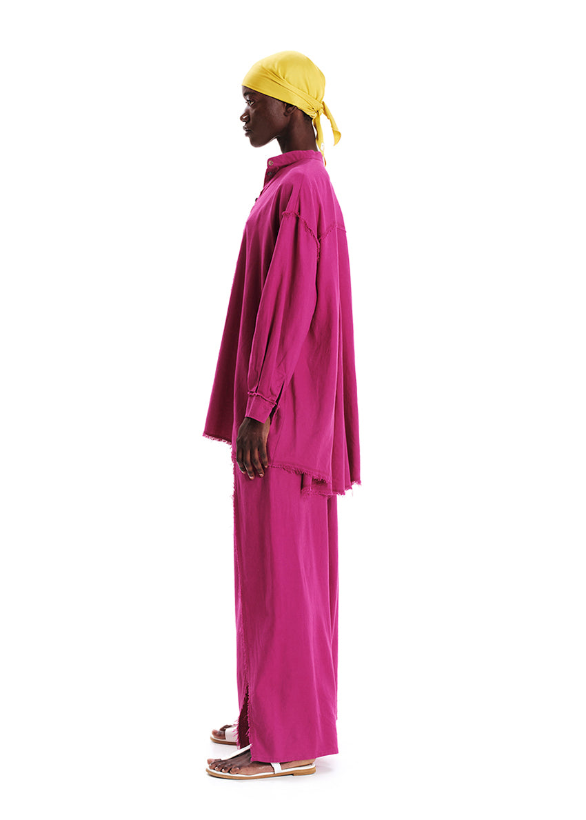A model wearing a magenta oversized linen and bamboo blend shirt with fringes at the seams, paired with matching pants.