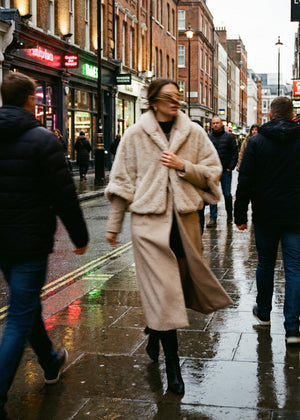 Woman in a beige coat walking on a city street with pedestrians around.