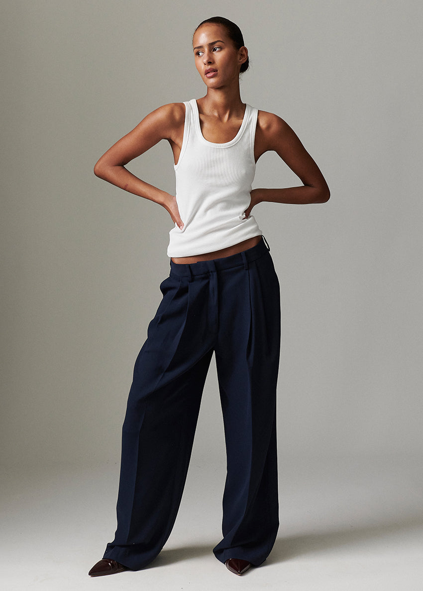 Woman wearing a white tank top and navy pants against a neutral background