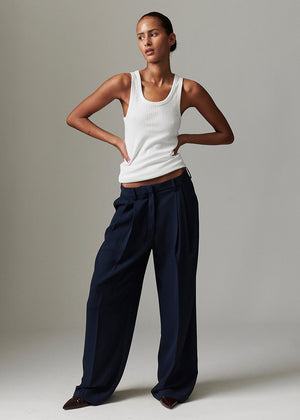 Woman wearing a white tank top and navy pants against a neutral background