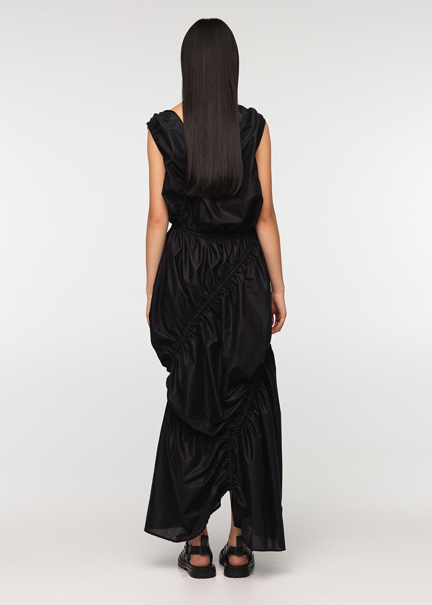 A long sleeveless asymmetrical black dress with a deep V neckline, featuring diagonal seams and gathering on all sides, modeled by a woman standing against a white background.