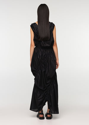 A long sleeveless asymmetrical black dress with a deep V neckline, featuring diagonal seams and gathering on all sides, modeled by a woman standing against a white background.