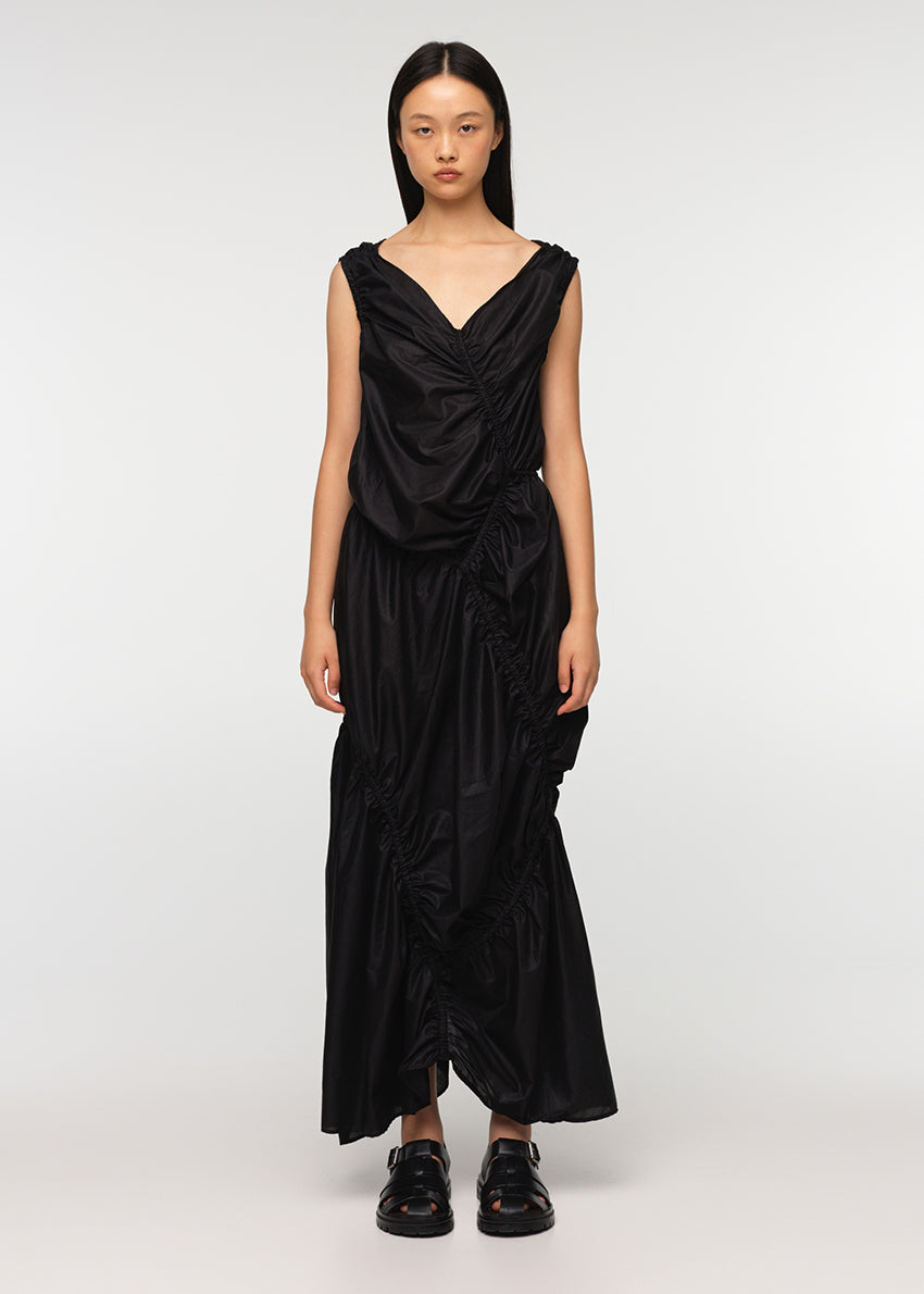 A long sleeveless asymmetrical black dress with a deep V neckline, featuring diagonal seams and gathering on all sides, modeled by a woman standing against a white background.