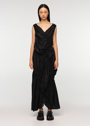 A long sleeveless asymmetrical black dress with a deep V neckline, featuring diagonal seams and gathering on all sides, modeled by a woman standing against a white background.