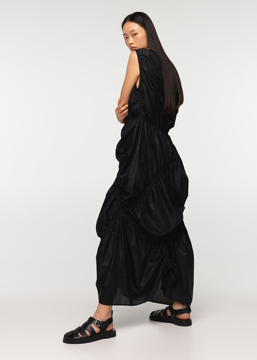 A long sleeveless asymmetrical black dress with a deep V neckline, featuring diagonal seams and gathering on all sides, modeled by a woman standing against a white background.