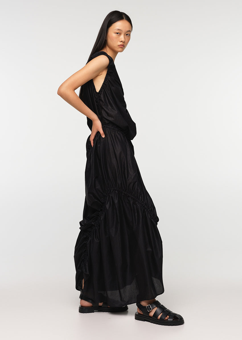 A long sleeveless asymmetrical black dress with a deep V neckline, featuring diagonal seams and gathering on all sides, modeled by a woman standing against a white background.
