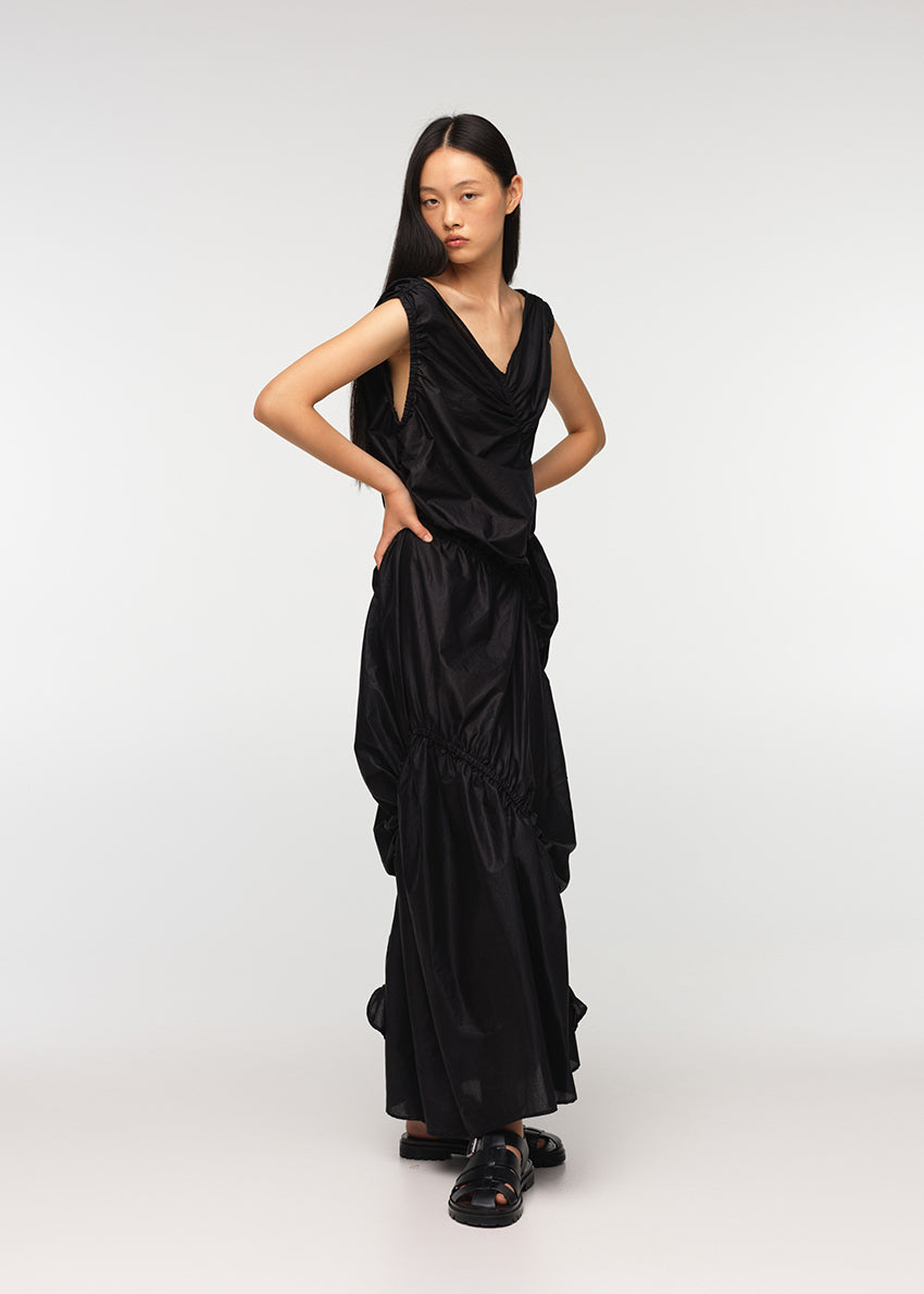 A long sleeveless asymmetrical black dress with a deep V neckline, featuring diagonal seams and gathering on all sides, modeled by a woman standing against a white background.