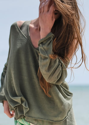 A woman with long, curly hair wearing a khaki green short sleeve pullover with a deep V-neckline, standing near the beach.