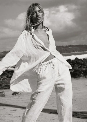 A person standing on a beach wearing white linen pants with a relaxed fit and an adjustable waistband, paired with a white shirt.