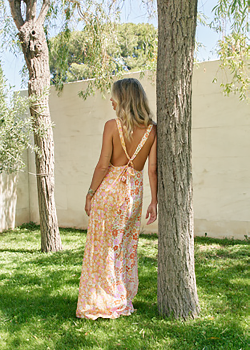 A woman wearing a sleeveless, floral print, maxi dress with a V-neckline and an open back standing on grass.