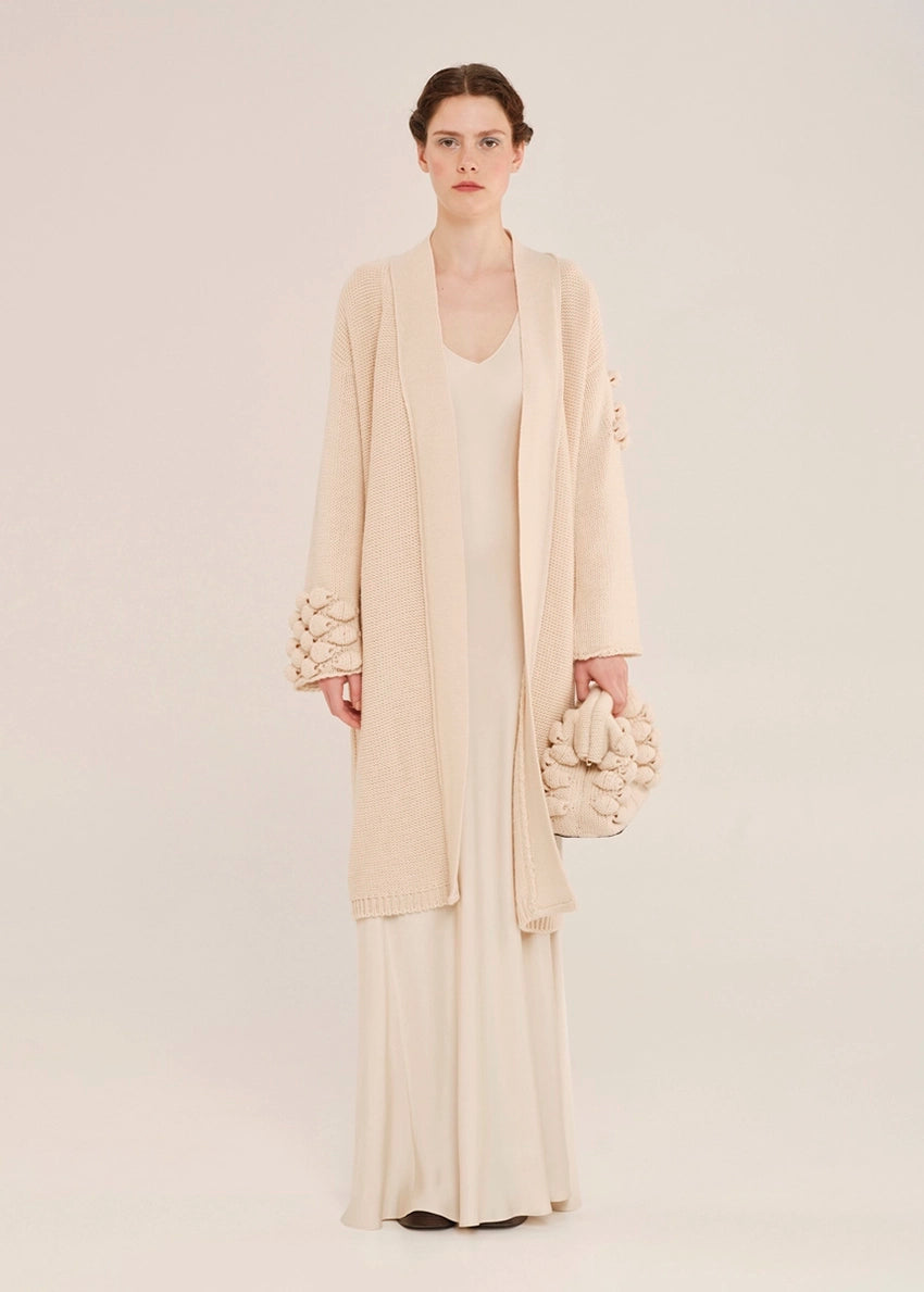 Woman wearing a long brown cardigan with textured cuffs and a matching scarf in an indoor setting.