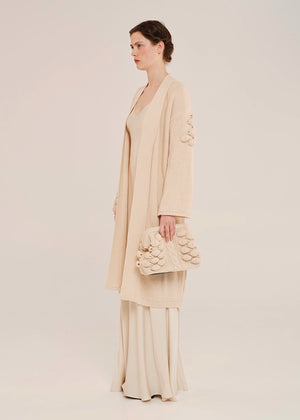 Woman in a beige outfit holding a matching handbag against a plain background