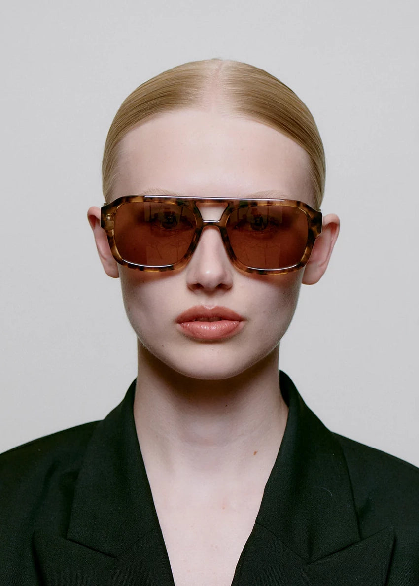 Person wearing a black outfit and brown sunglasses against a plain background