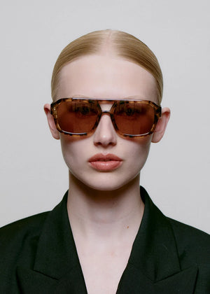 Person wearing a black outfit and brown sunglasses against a plain background