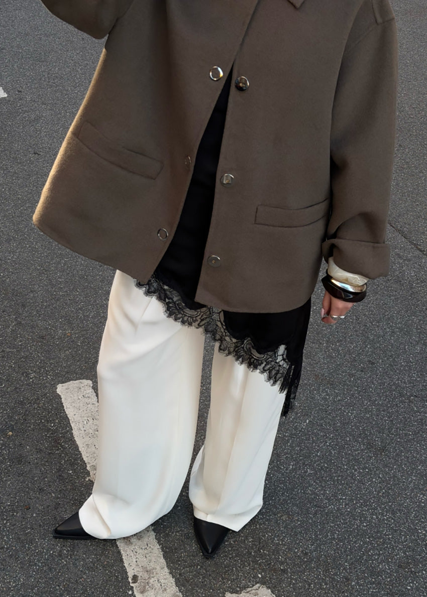 Person wearing a brown coat, white pants with lace trim, and black shoes on a gray pavement.
