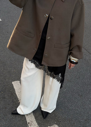 Person wearing a brown coat, white pants with lace trim, and black shoes on a gray pavement.