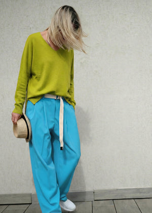 Person wearing a green sweater and blue pants against a neutral wall.