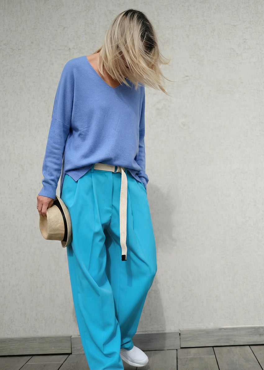 Person wearing a blue sweater and turquoise pants holding a hat against a neutral background