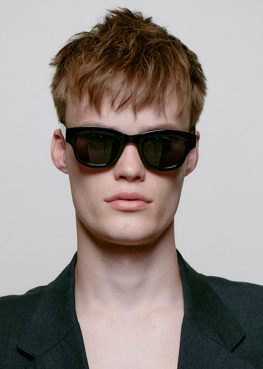 Person wearing black sunglasses and a dark jacket against a plain background