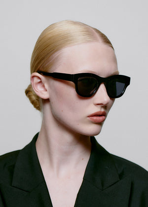 Person wearing black sunglasses and a black outfit against a plain background
