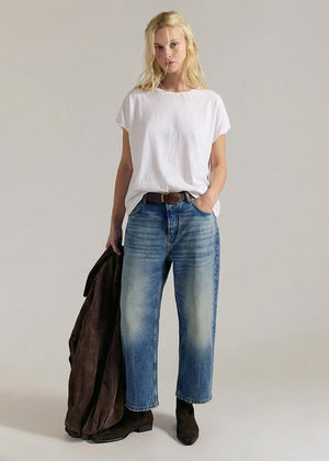 Woman wearing a white t-shirt and blue jeans holding a brown jacket on a plain background