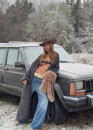 Person in cowboy hat and cardigan leaning against a vintage car in a snowy forest.