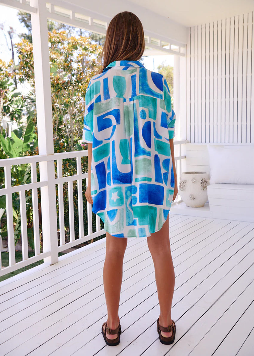 A woman wearing an oversized, geometric-patterned shirt in blue and green colors, with long sleeves and a collar, posing outdoors.