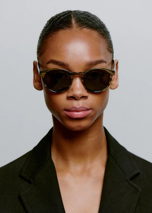 Person wearing dark sunglasses and a black blazer against a plain background