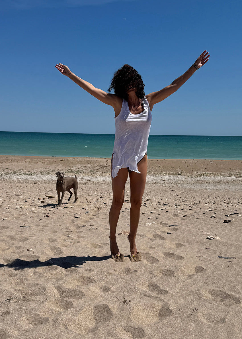Woman in white top with arms outstretched on a beach with a dog nearby