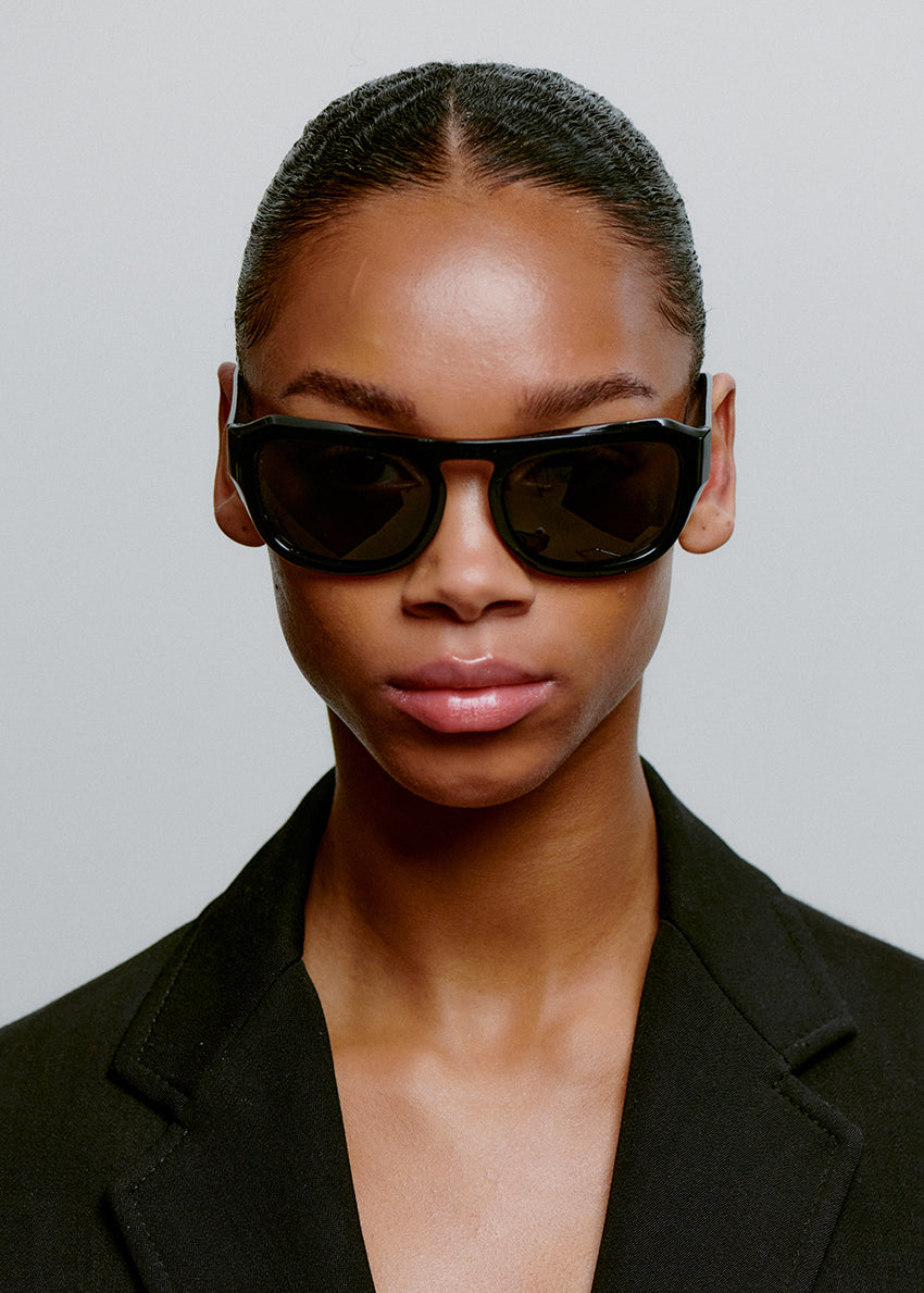 Person wearing black sunglasses and a black outfit against a gray background