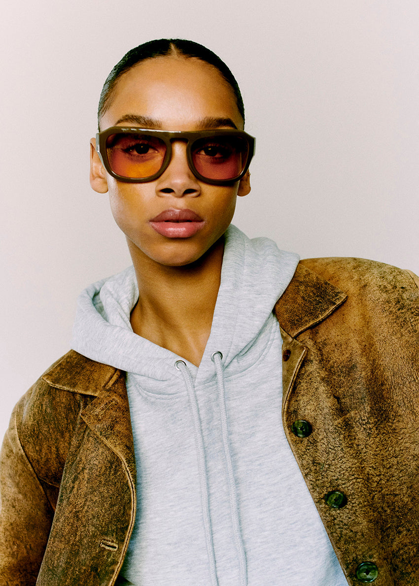 Person wearing sunglasses and a brown jacket over a gray hoodie against a plain background